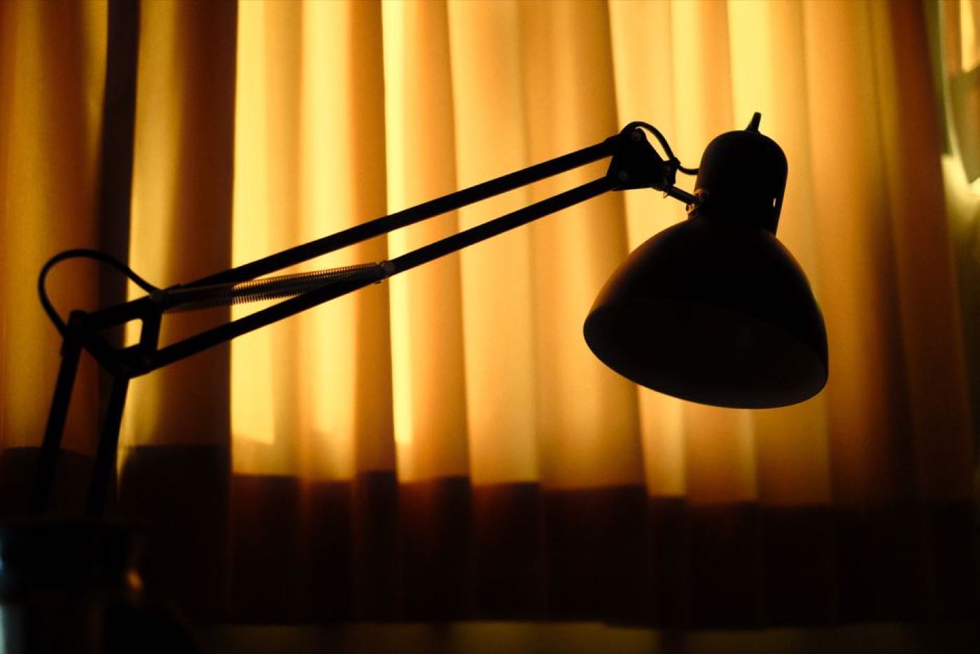 An angled desk lamp silhouetted against warm backlit curtains in a dim room.