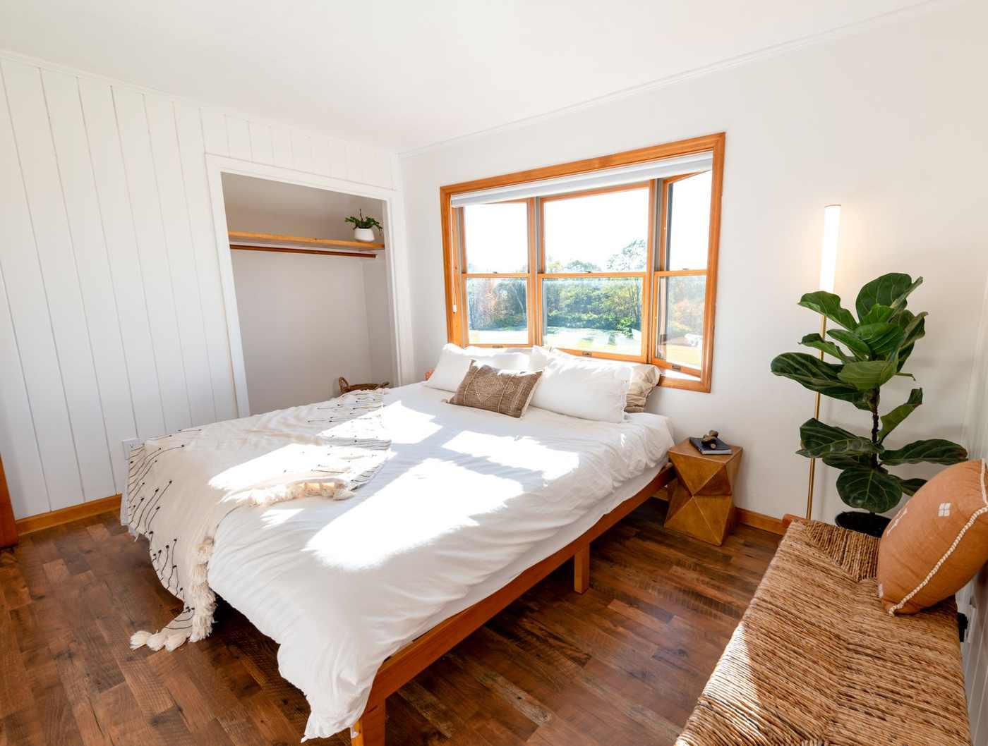 A bright bedroom with white bedding and a wooden floor, with strong summer sunlight falling across the bed from a large window.