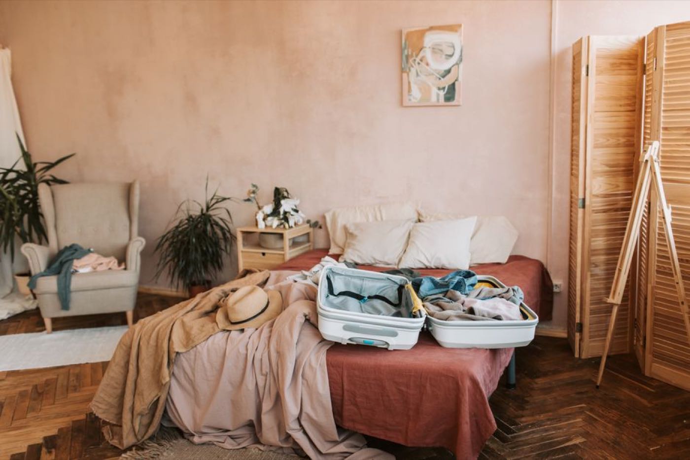 An open suitcase part-packed on a bed in a warm bedroom, with clothes and a sun hat beside it.