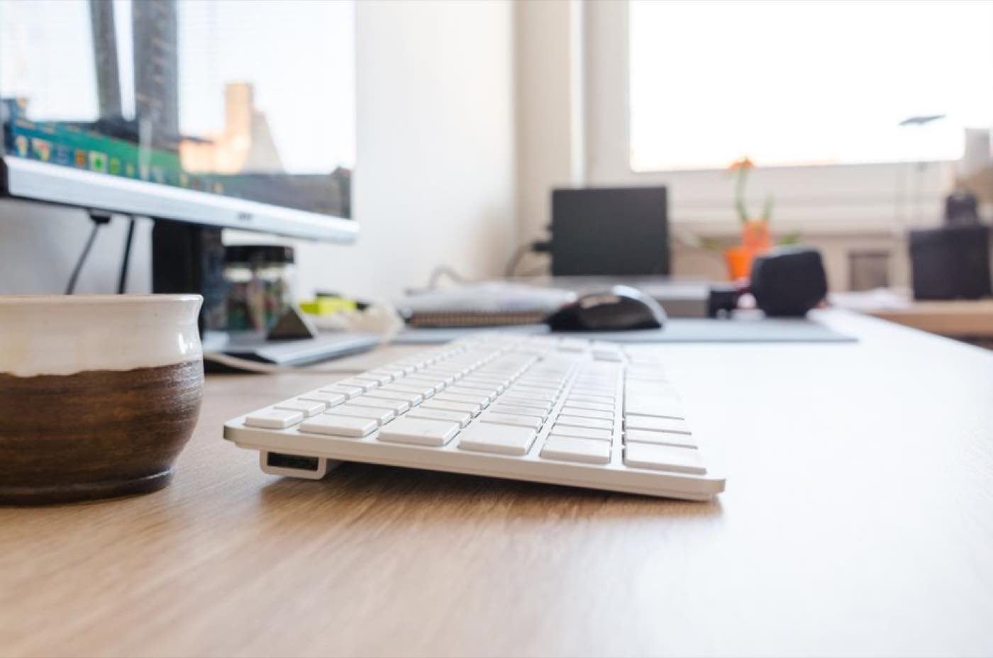 A keyboard and coffee mug on a wooden desk beside a bright window.